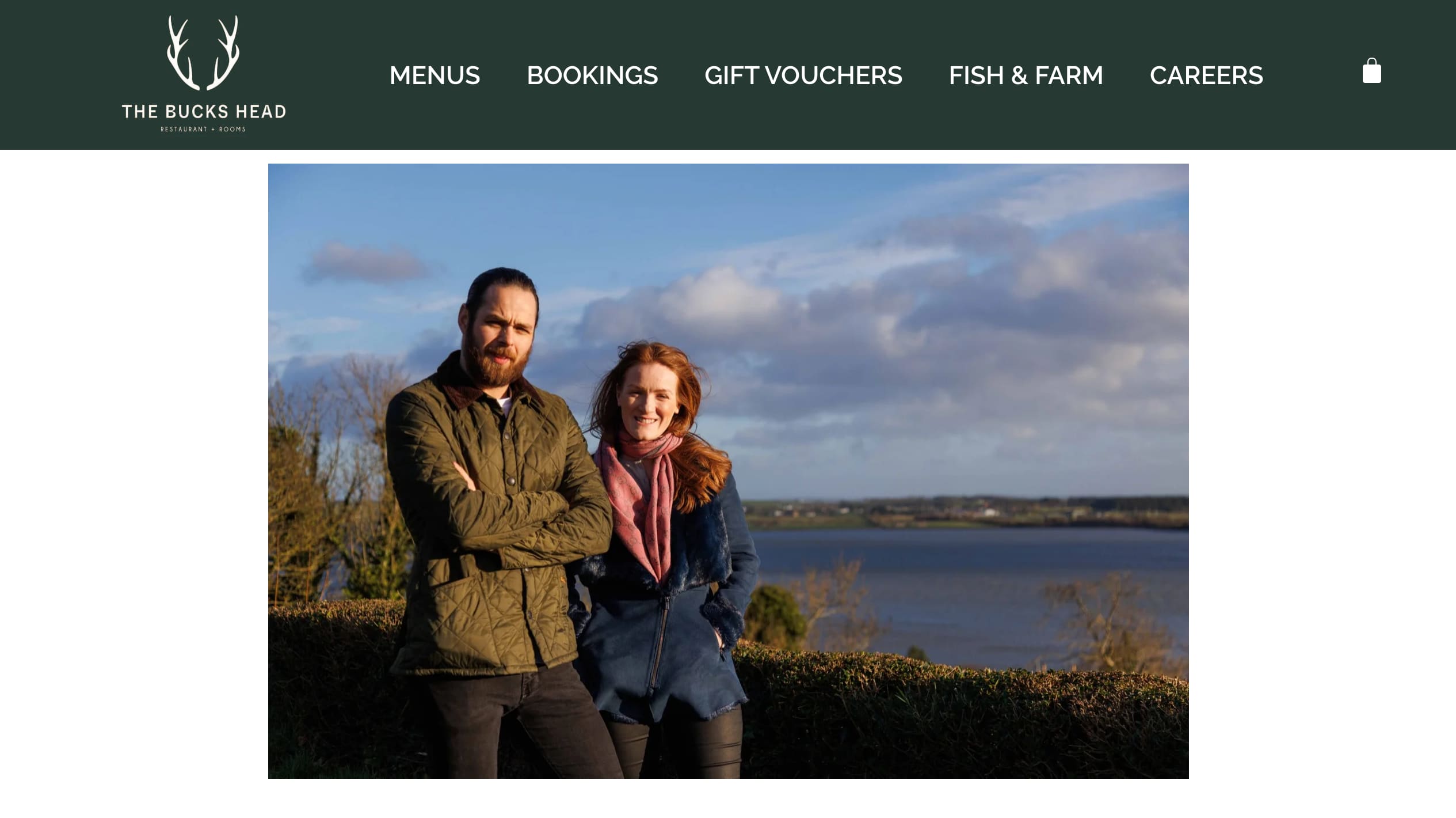 The Bucks Head's current homepage: a green header with the antler logo and five menu items above a full-width photograph of the owners by Dundrum Bay, with no headline, hours or booking action