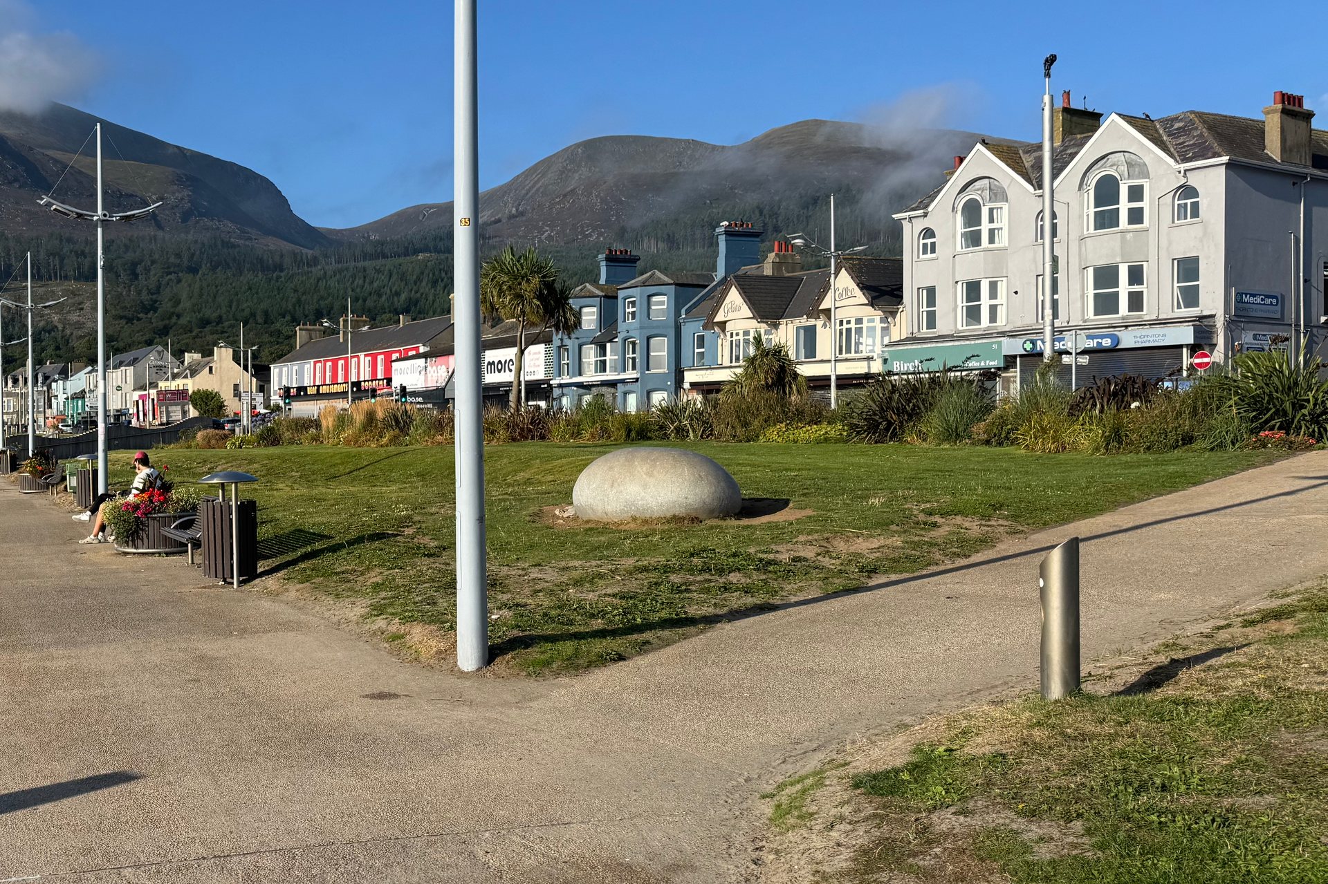 The Central Promenade in Newcastle on a sunny morning: a row of painted shopfronts behind a green with a large stone sculpture, and Slieve Donard rising behind them with cloud on its summit.